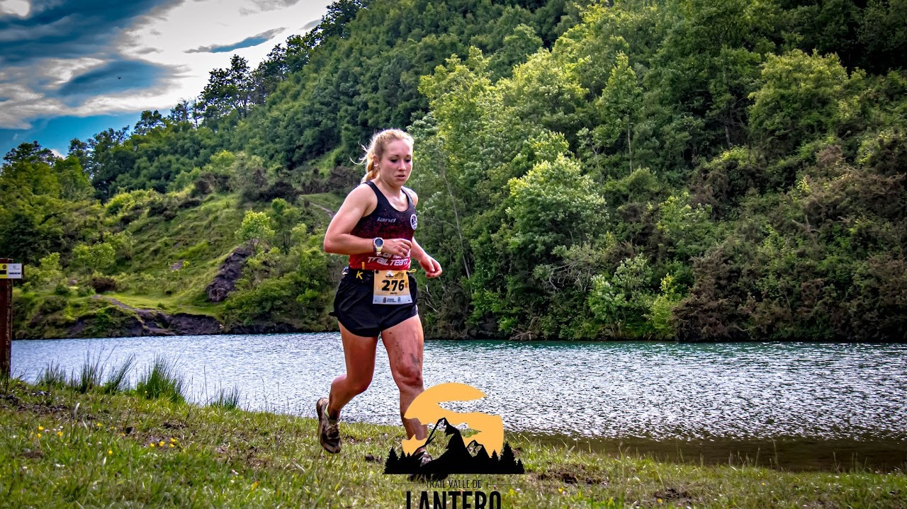 Corredora con dorsal 276 corre por la orilla de una laguna con el bosque detrás