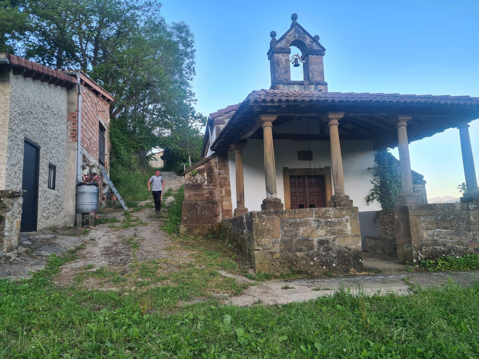 Capilla de Villacedré, con su pórtico de columnas y la espadaña, junto a una casa de piedra